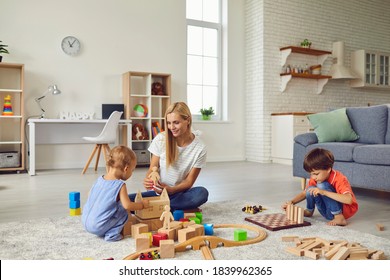 Family At Home. Happy Mother Playing With Little Children Of Different Ages In Cozy Studio Apartment. Babysitter And 2 And 5 Year Old Kids Building Wood Block Town On Warm Floor In Living Room