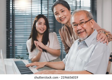 Family Home Health Care Wife And Daughter Caregiver Take Care Senior Elderly Retirement Father At Home Sitting On The Wheel Chair Playing Piano Together.