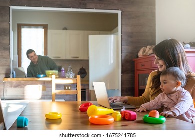 Family At Home In The Kitchen, Mom Works At The Computer At The Table With Her Daughter On Her Legs While Playing With Toys, The Father Cooks Lunch - Millennials In An Intimate Moment With Their Child