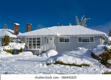 Family House At Winter Season. Residential House In Snow On A Sunny Day. Grey House In Snow On Blue Sky Background.