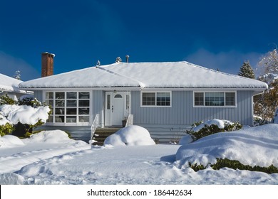 Family House At Winter Season. Residential House In Snow On A Sunny Day. Grey House In Snow On Blue Sky Background.