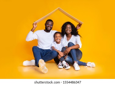 Family Housing Concept. Happy African American Parents And Their Daughter Sitting Under Symbolic Roof Dreaming About New Home, Yellow Background