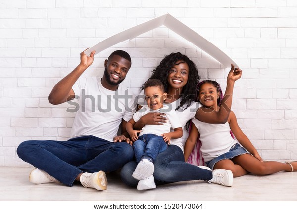 Family Housing Concept. Young African American\
Parents With Two Children Sitting Under Cartboard Roof Dreaming Of\
New Home, Copy Space