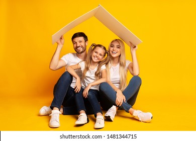 Family Housing Concept. Young Parents And Daughter Sitting Under Roof Dreaming Of New Home. Yellow Studio Background, Copy Space