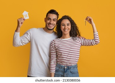 Family Housing. Happy Arab Couple Holding Home Keys And Paper House Icon, Joyful Middle Eastern Spouses Celebrating Moving To Their Own Apartment, Standing Over Yellow Studio Background, Free Space