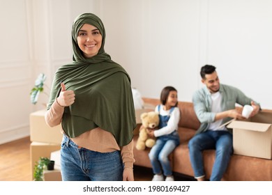 Family Housing. Portrait Of Cheerful Muslim Woman In Hijab Showing Thumbs Up Gesture, Arabic Family Celebrating Their Moving Day, Dad And Daughter Sitting On Couch In New House Unpacking Boxes