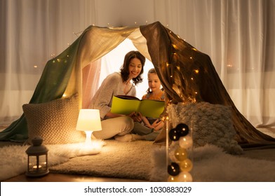 Family, Hygge And People Concept - Happy Mother And Little Daughter Reading Book In Kids Tent At Night At Home