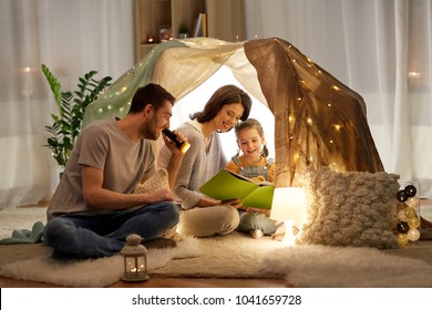 family, hygge and people concept - happy mother, father and little daughter reading book with torch light in kids tent at night at home - Powered by Shutterstock