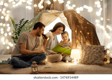 Family, Hygge And People Concept - Happy Mother, Father And Little Daughter Reading Book With Torch Light In Kids Tent At Night At Home