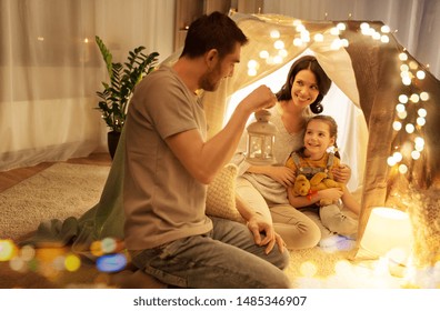 Family, Hygge And People Concept - Happy Mother, Father With Lantern And Little Daughter With Teddy Bear Playing In Kids Tent At Night At Home