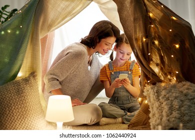 Family, Hygge And Technology Concept - Happy Mother And Little Daughter With Smartphone In Kids Tent At Night At Home