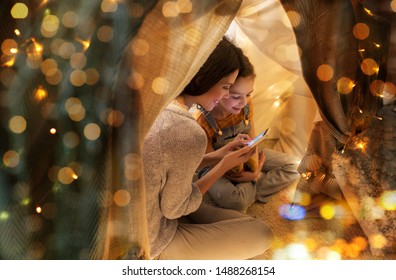 Family, Hygge And Technology Concept - Happy Mother And Little Daughter With Smartphone In Kids Tent At Night At Home