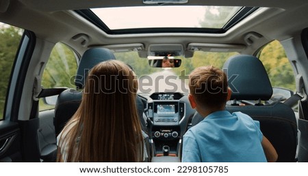 Family idyll concept where lovely happy brother and sister sitting at the rear seat of the auto,watch the way they are driving on and tell stories to their mom who's behind the wheel