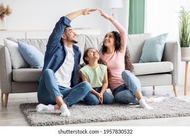 Family Insurance Concept. Young Arab Parents Making Roof Symbol Of Hands Above Their Little Daughter While Sitting On Floor At Home, Middle Eastern Mom, Dad And Child Enjoying New House, Free Space - Shutterstock ID 1957830532