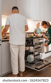 Una familia interactúa en su cocina moderna y luminosa. El niño se sienta en el mostrador cerca del lavavajillas abierto mientras los padres se encargan de las tareas domésticas. La escena muestra la vida diaria y el trabajo en equipo en casa.