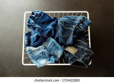 Family Jeans In Laundry (washing) Basket On Black Chalkboard Background. Denim Trousers Captured From Above (top View). Various Sizes, Shapes And Blue Shades (tones).