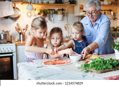 Family, Kids Cooking Pizza In Cozy Home Kitchen. Grandmother And Three Sisters, Her Granddaughters Preparing Homemade Italian Food. Funny Little Girls Are Helping Senior Woman. Children Chef Concept.