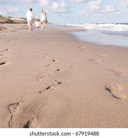 Family Leaves Footprints In The Sand As Parents Walk With Baby On The Beach