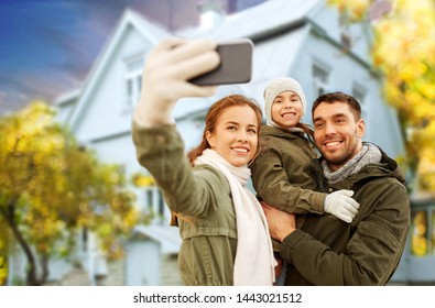 Family, Leisure And People Concept - Happy Mother, Father And Little Daughter Taking Selfie By Smartphone Over House In Autumn Background