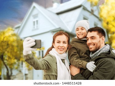 Family, Leisure And People Concept - Happy Mother, Father And Little Daughter Taking Selfie By Smartphone Over House In Autumn Background