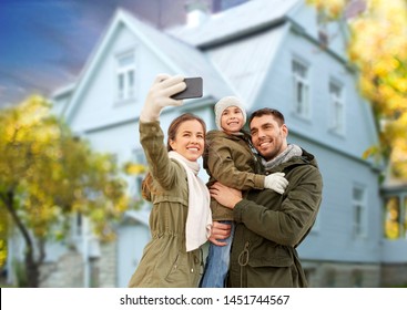 Family, Leisure And People Concept - Happy Mother, Father And Little Daughter Taking Selfie By Smartphone Over House In Autumn Background