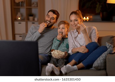 family, leisure and people concept - happy smiling father, mother and little son eating popcorn and watching tv at home in evening - Shutterstock ID 1635301450