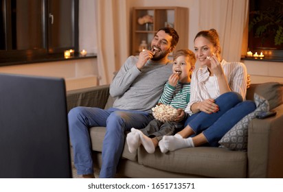 family, leisure and people concept - happy smiling father, mother and little son watching tv and eating popcorn at home at night
