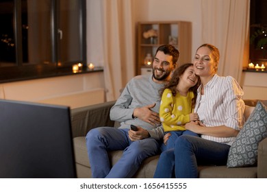 family, leisure and people concept - happy smiling father, mother and little daughter watching something funny on tv at home at night