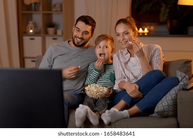 concepto de familia, ocio y gente - padre, madre y hijo sonrientes felices comiendo palomitas de maíz y viendo televisión en casa por la noche
