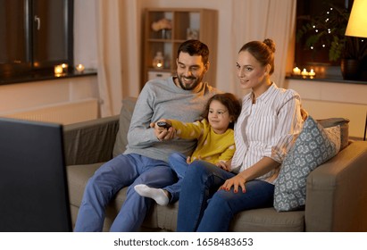 Family, Leisure And People Concept - Happy Smiling Father, Mother And Little Daughter With Remote Control Watching Tv At Home At Night