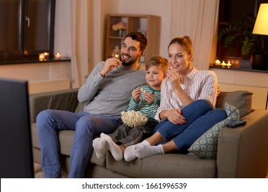 family, leisure and people concept - happy smiling father, mother and little son watching tv and eating popcorn at home at night