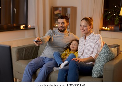 family, leisure and people concept - happy smiling father with remote control, mother and little daughter watching tv at home at night