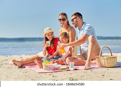 family, leisure and people concept - happy mother, father and two daughters having picnic on summer beach and drinking juice