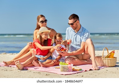 family, leisure and people concept - happy mother, father and two daughters having picnic on summer beach and eating strawberries