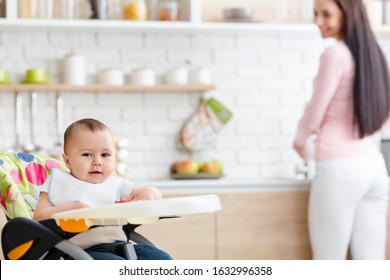 Family Life. Baby Playing On High Chair In Kitchen, Mother Washing Dishes On Background, Empty Space