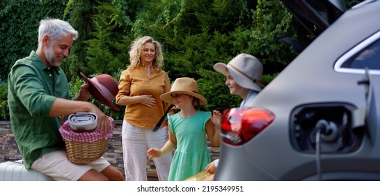 Family With Little Children Loading Car And Waiting For Charging Car Before Going On Picnic.