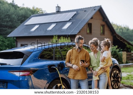 Family with little girl standing in front of their house with solar panels on the roof, having electric car.