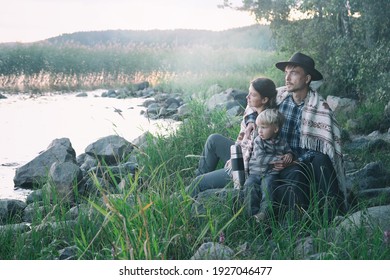 Family Of Local Travellers Father Mother And Child Wrapped In Whool Blanket Sitting Near Lake On Sunset. Weekend Picnic On Nature, Travel With Kids, Staycation, Folk Lifestyle.