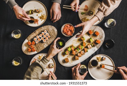 Family lockdown Japanese sushi dinner from delivery service at home. Flat-lay of table with salmon, crab, prawn, vegan rolls, wasabi, ginger and people eating together over dark background, top view