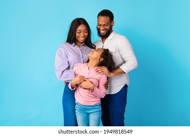 Family Love Concept. Portrait Of African American Man And Woman Hugging And Looking At Smiling Curly Girl Standing Isolated On Blue Studio Background. Mum And Dad Posing With Daughter At Camera