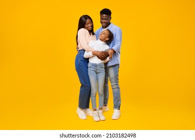 Family Love Concept. Portrait Of African American Man And Woman Hugging And Looking At Smiling Girl Standing Isolated On Yellow Studio Background. Mum And Dad Posing With Daughter, Full Body Length