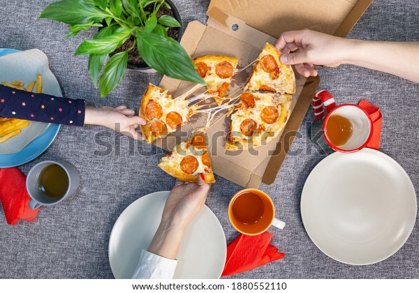 Family lunch or dinner with pepperoni pizza.\
One child\'s and two adults\' hands take slices of mini pizza with\
sausage from a craft box, top view. Food delivery, ordering food at\
home, pizzeria.