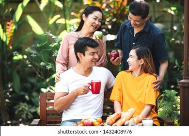 Family Lunch Outdoor. Married Mix Race Couple With Grand Parents. White Man, Asian Pregnant Woman With Senior Asian Couple In Tropical Garden. Talking To Eath Other, Laughing.