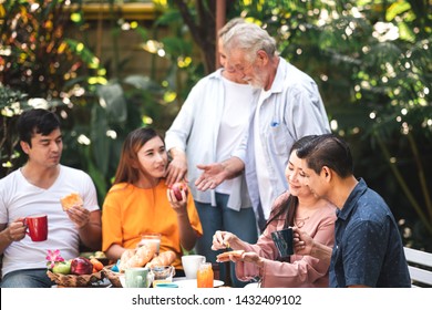 Family Lunch Outdoor. Married Mix Race Couple With Grand Parents. White Man, Asian Pregnant Woman With Senior Asian, White Couple In Tropical Garden. Eating Bread.