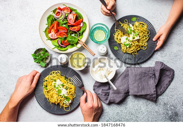 Family lunch table, top view. Peoples hands\
eating italian pasta with ricotta and fresh vegetables salad.\
Italian cuisine concept.