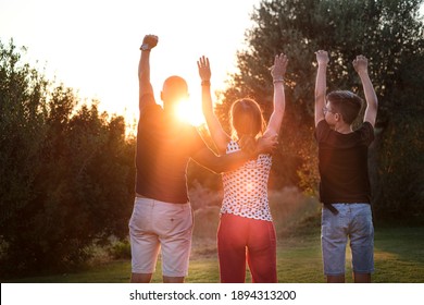 Family Made Up Of Three People, From Behind, Hugging Each Other As A Sign Of Union Facing The Sunset In A Natural Context