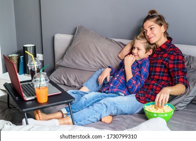 Family Of Mama And Son Watching Movie Online In Laptop With Eating Popcorns On Messy Bed At Home. Quarantine. Stay At Home. Sport Equipment For Outdoor Activitieson The Bed. Coronavirus Situation.