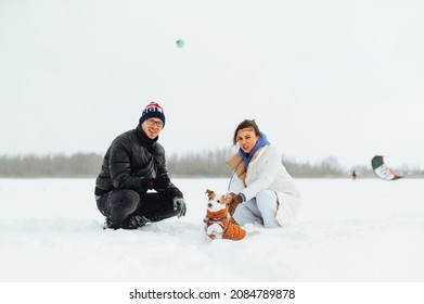 Family Man And Woman Playing With A Cute Dog In A Jack Russell In The Snow With The Help Of A Ball With A Smile On His Face. Winter Games In The Snow On A Walk With A Pet.