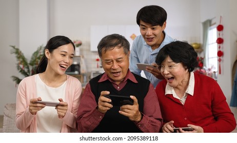 Family Members Having Fun Playing Games On Smartphone During Chinese New Year Holiday At Home. Everyone Leaning Close To Happy Old Father Who Just Win The Game