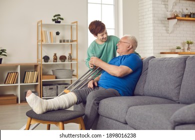 Family members take good care of senior man with broken leg. Little child helps injured grandfather during recovery period. Grandson giving glass of water to grandpa sitting on sofa with crutches